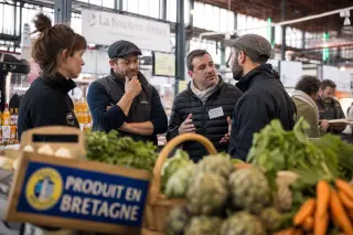 Regional shopkeepers and fair organizers in a Rennes market hall sharing a natural conversation amid stalls with local products, softly lit by morning light.