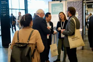 Businesspeople and local partners interact casually with coffee in hand in a lively corridor at Rennes Parc Expo, natural light creating an authentic, relaxed atmosphere.
