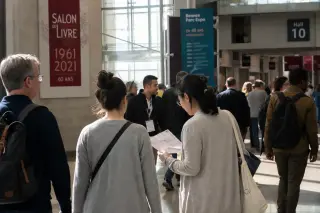 Visitors of various ages and backgrounds walk and talk in a sunlit hallway at the Rennes Exhibition Center, captured candidly in a documentary style.