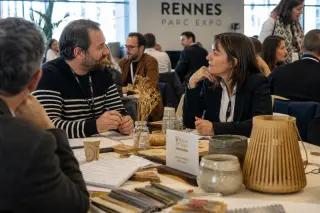 Artisans and professionals in a softly lit conference room at Rennes exhibition center, engaged in quiet, natural discussion around a table with craft samples and coffee cups.