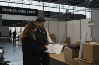 Scene inside Rennes exhibition center showing two entrepreneurs at an innovative stand, visitors interacting with displays, soft daylight and authentic exhibition bustle.