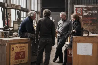 Several mature workers casually converse near old fair equipment and crates backstage in Rennes' exhibition hall, surrounded by quiet activity and natural daylight.
